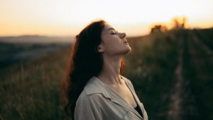 Woman standing with eyes closed in a field during sunset, exuding tranquility and peace.